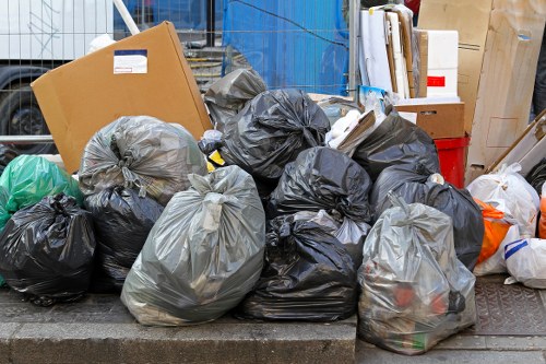 A collection of black, grey, and clear rubbish bags filled with waste materials, placed on a pavement or driveway area in an outdoor setting. The bags are made of plastic, some appearing slightly shiny and crinkled, with various sizes and shapes, indicating different types of discarded rubbish. Behind the bags, there are partially visible cardboard boxes, a red container, and other miscellaneous items, suggesting a waste clearance or rubbish removal process near a residential or commercial property in West Wickham. The setting appears to be an urban area with a textured ground surface, possibly concrete or asphalt, and the natural lighting indicates daytime conditions. The scene is typical of a rubbish collection or house clearance activity, which a company like House Clearance West Wickham might handle, focusing on outdoor waste disposal in the local area.