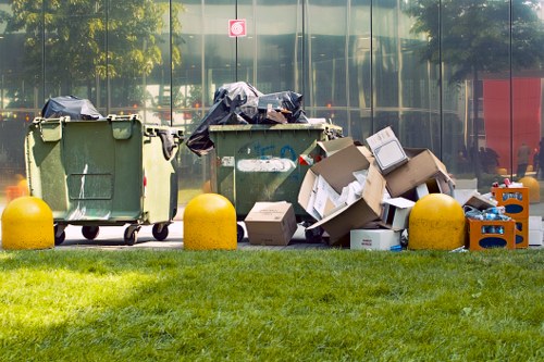 A row of three large green waste bins and several overflowing cardboard boxes placed on the pavement next to a grassy area, with yellow wheel stops in front of the bins. The cardboard boxes contain various discarded items, some torn or crumpled, and are partially open or flattened, indicating recent disposal. Black rubbish bags are draped over the top of two of the bins, which are positioned in front of a modern glass building reflecting trees and vehicles on the street. The scene suggests a clearance or rubbish removal operation outside a property, possibly in a residential or commercial area within West Wickham, highlighting the need for professional rubbish removal services offered by House Clearance West Wickham.