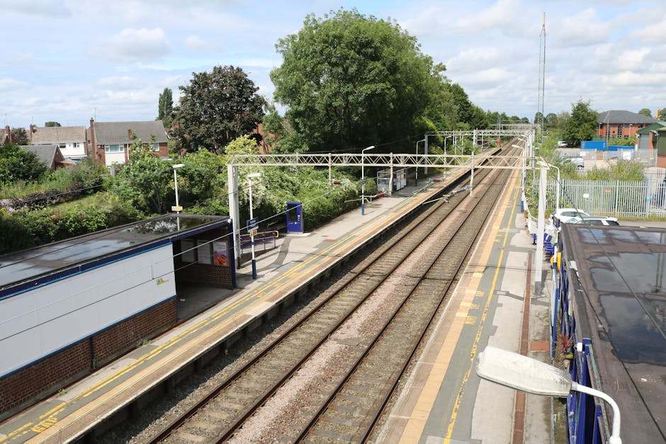 An elevated view of a railway station platform on a clear day, featuring parallel train tracks with gravel and wooden sleepers, bordered by a paved waiting area with tactile paving for visually impair